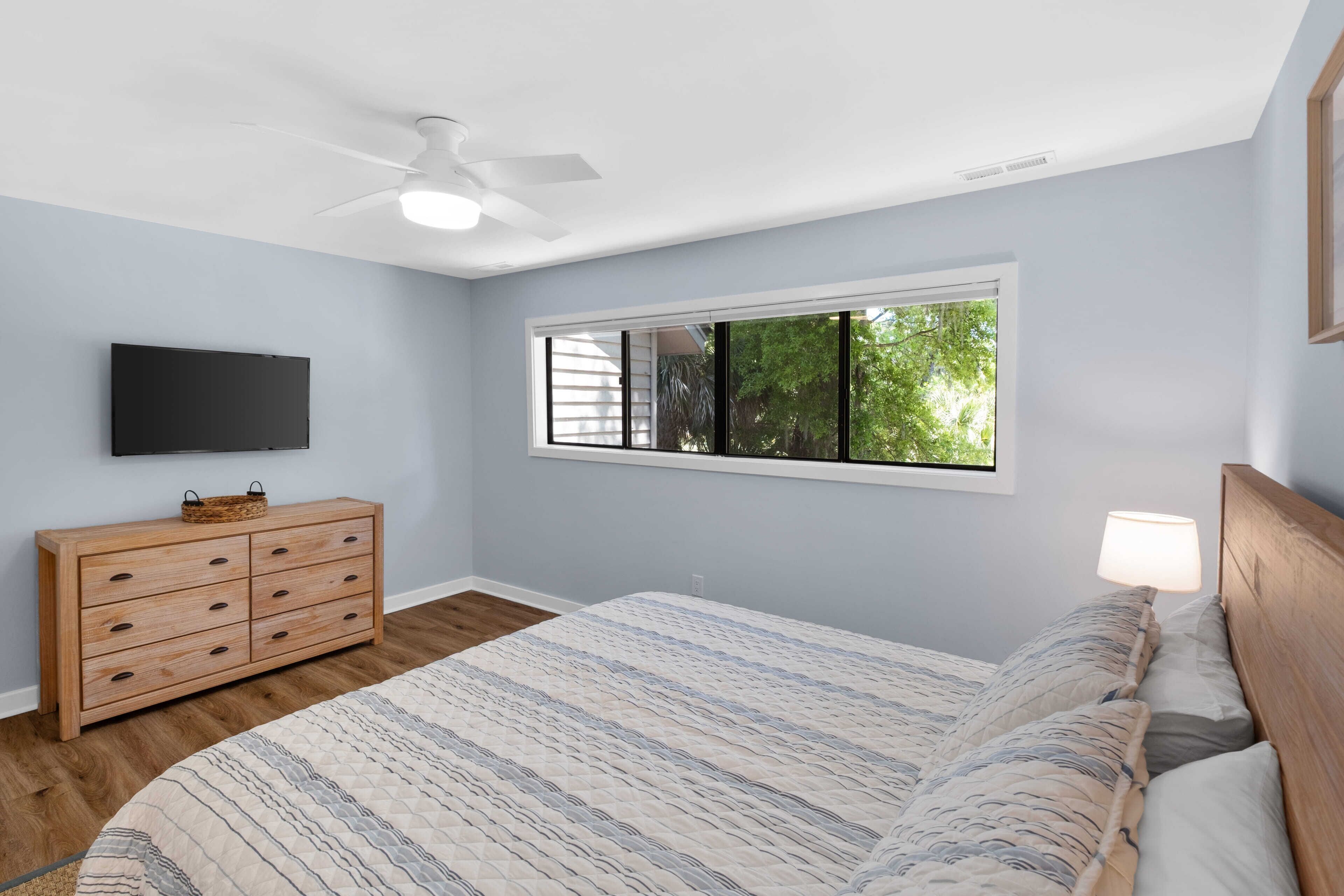 A bedroom with a bed, a dresser, a television, and a window with a view of trees.