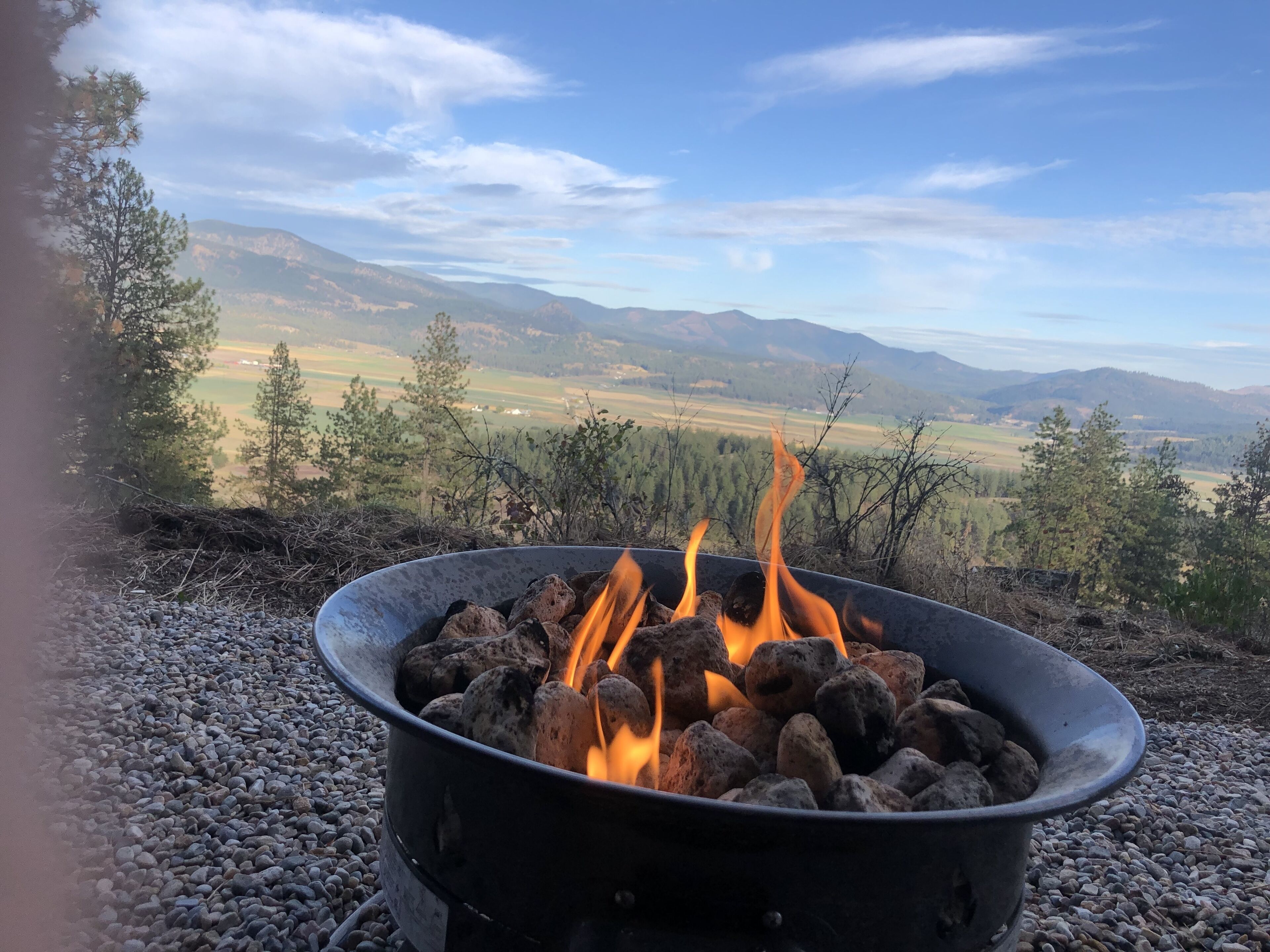 A campfire in a metal bowl on a gravel surface with a scenic view of mountains and trees in the background.