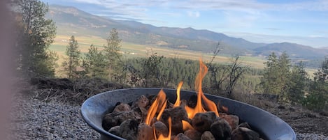 A campfire in a metal bowl on a gravel surface with a scenic view of mountains and trees in the background.