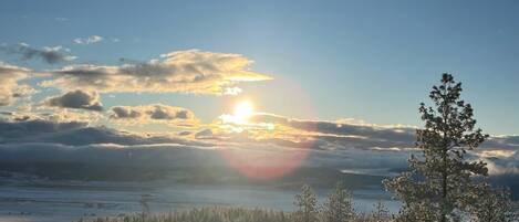 A landscape with a sun setting behind clouds, a snow-covered field, and a forest of trees.