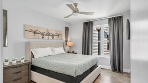A bedroom with a bed, a nightstand, a ceiling fan, a window with curtains, and a wall-mounted artwork.