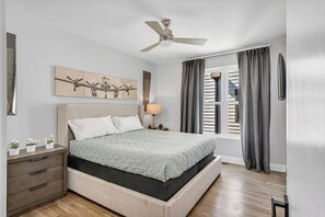 Ein Schlafzimmer mit Bett, Nachttisch, Deckenventilator, Fenster mit Vorhängen und Wandkunst.