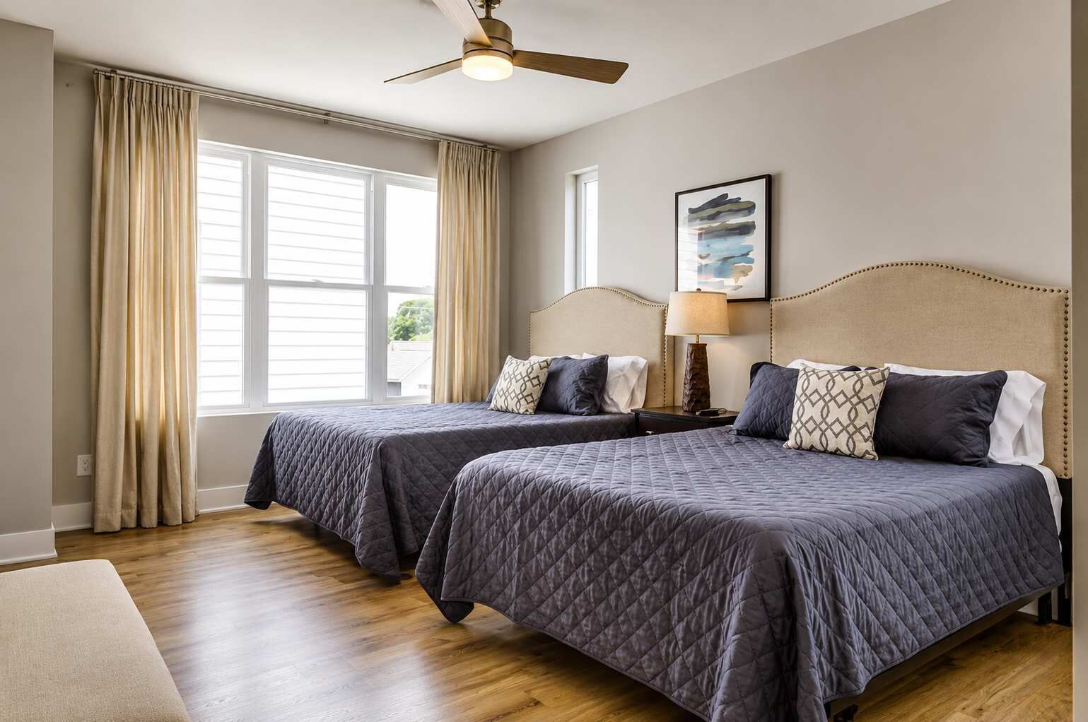 Habitación de hotel con dos camas tamaño queen, ventilador de techo, ventana con cortinas e imagen enmarcada en la pared.