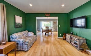 A living room with a plaid sofa, a dining table with chairs, a TV mounted on the wall, and a bookshelf. - 2 blocks to Olympic Oval, Walk to Downtown, Mountain Views! (Lake Placid)