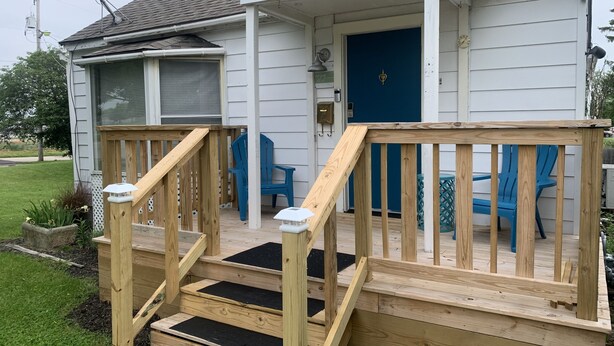 Una terraza de madera con una rampa que lleva a una puerta azul, rodeada de sillas azules.