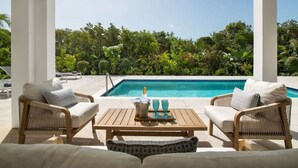 Zona junto a la piscina con mobiliario de mimbre, una mesa con una botella y vasos, y vistas a una piscina rodeada de exuberante vegetación.