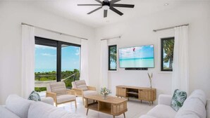 Una sala de estar moderna con un televisor grande, sofás blancos, una mesa de centro de madera y un balcón con vistas al océano.