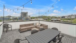 A rooftop terrace with a wooden table and wicker sofa, string lights, and a view of surrounding buildings. - Nashville Cowgirl Stay – Rooftop Views (Nashville)