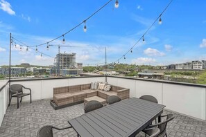 Una terraza en la azotea con mesa de madera, sofá de mimbre, luces colgantes y vistas de edificios circundantes.