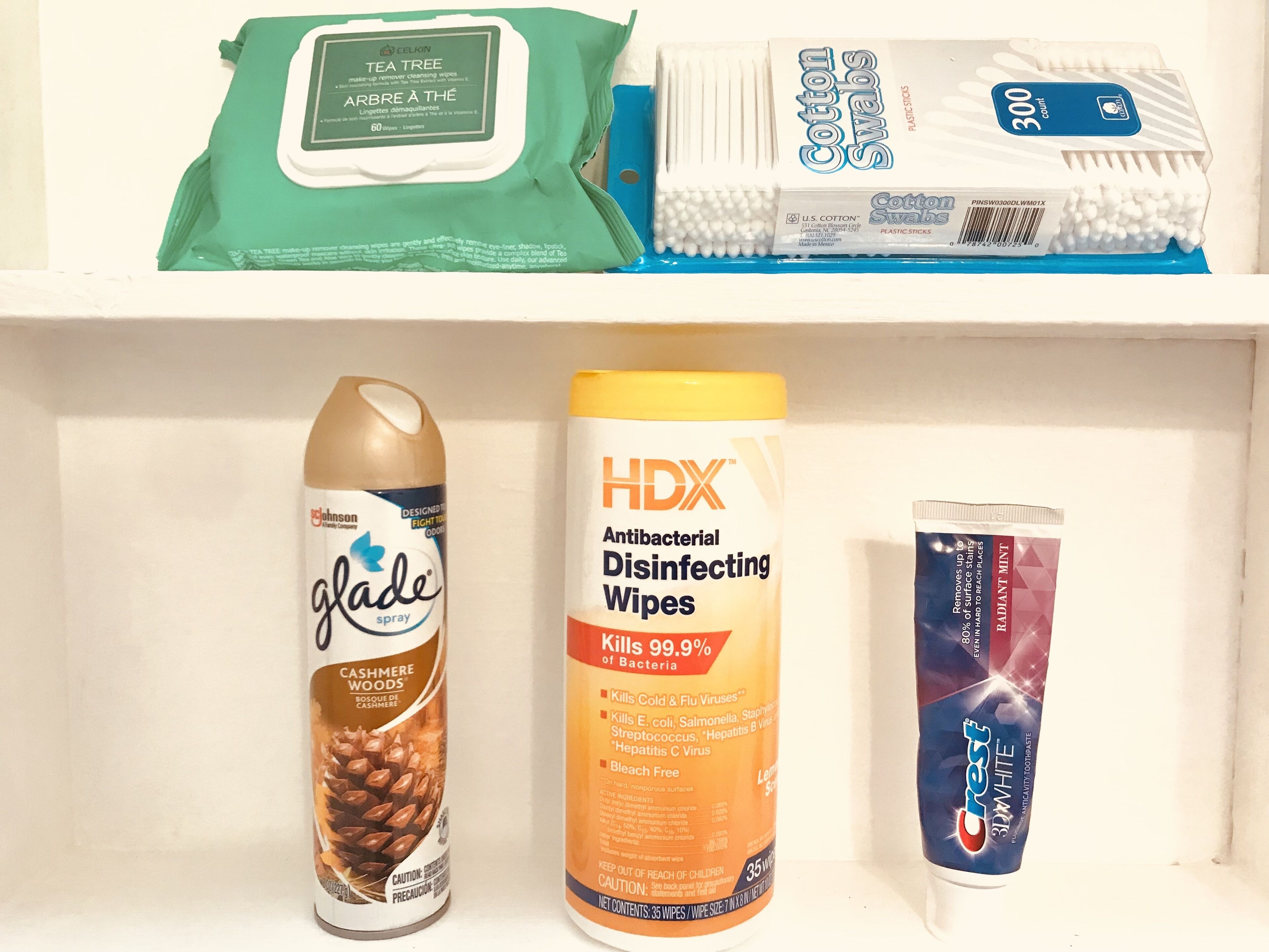 En hylla med olika rengöringsmedel, inklusive ett paket med rengöringsservetter med doft av grönt te och tea tree-olja, bomullspinnar, en sprayflaska märkt med texten ”glade”, en stor behållare med HDX-desinfektionsservetter och ett tub med Crest-tandkräm.