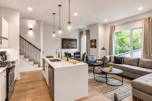 A modern kitchen with a central island, a dining area with a sofa and coffee table, and a living room with a large window.