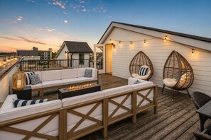 Terraza en la azotea con un sofá blanco, dos sillones de mimbre, una chimenea y luces colgantes.