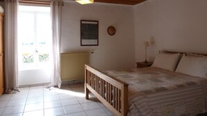 A bedroom with a wooden bed, a radiator, a hanging light fixture, a framed picture on the wall, and a window with curtains. - Gite1 T3 with swimming pool in the heart of the vineyards (34310 - CAPESTANG)