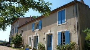 A two-story building with blue shutters, a gravel driveway, and potted plants. - Gite1 T3 with swimming pool in the heart of the vineyards (34310 - CAPESTANG)