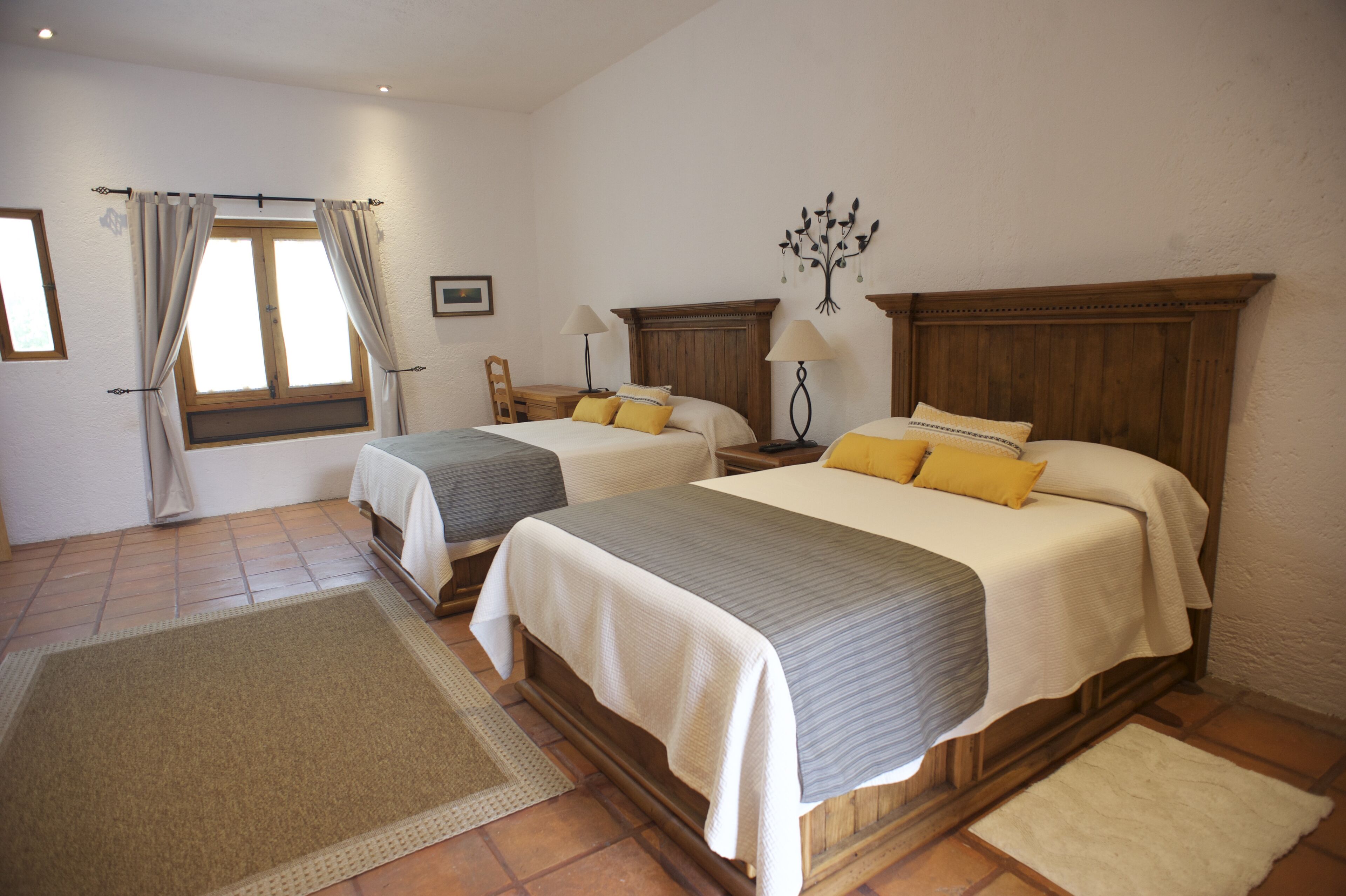 Une chambre d’hôtel avec deux lits, une tête de lit en bois, une table de chevet, une lampe, une fenêtre avec des rideaux, un tapis et un élément décoratif mural.