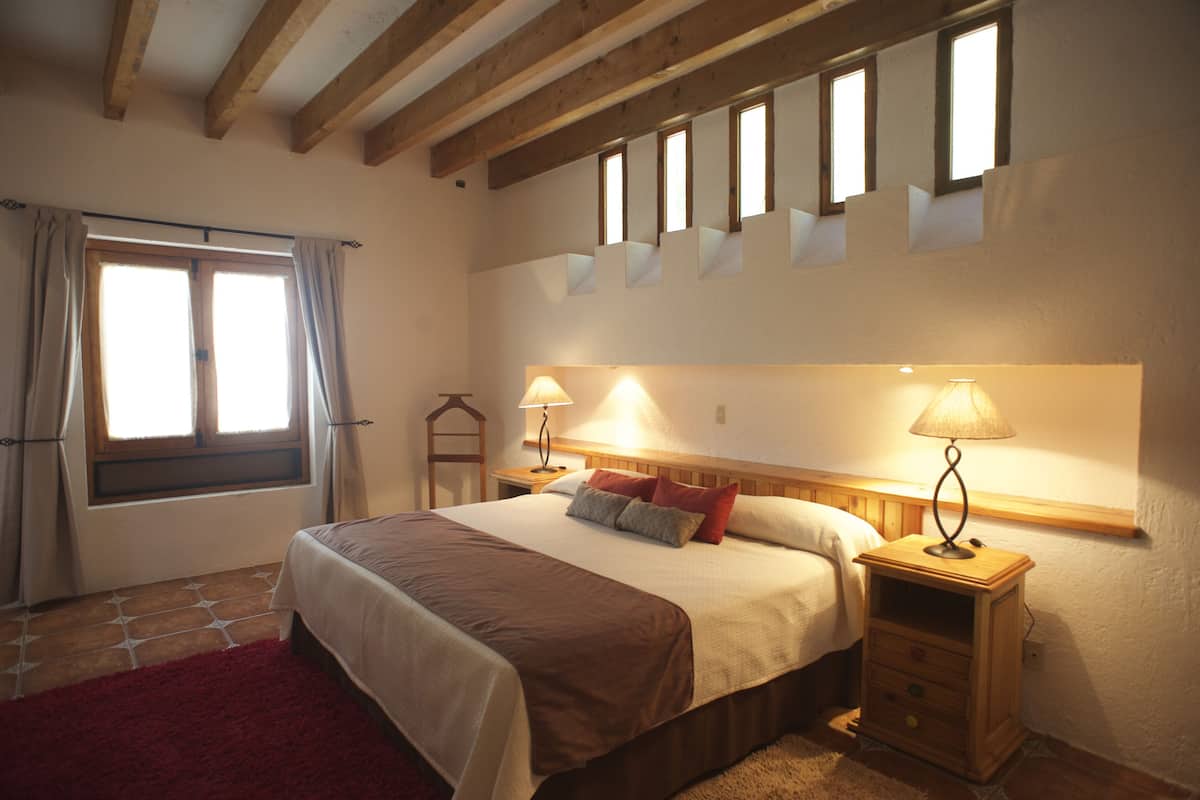 A bedroom with a large bed, two bedside tables with lamps, a window with curtains, and a wooden ceiling.