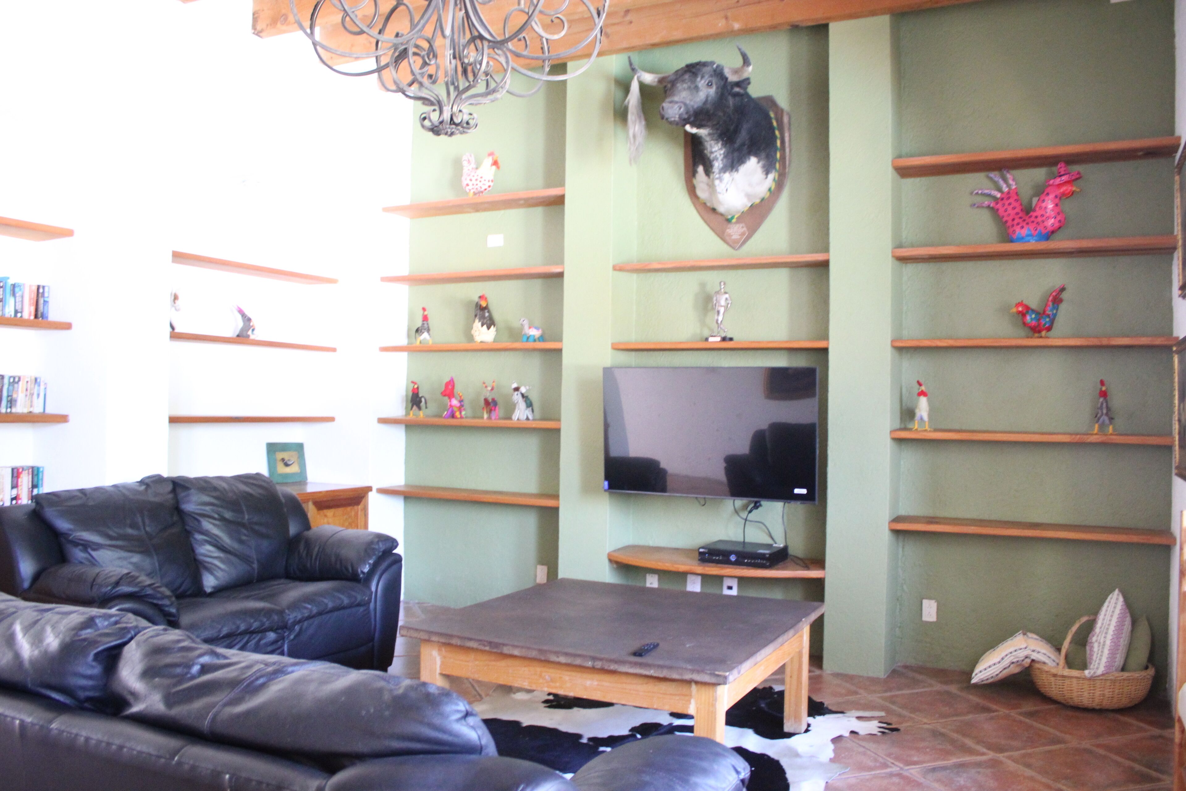 Un salon comprenant un canapé en cuir, une table basse en bois, un téléviseur fixé au mur et des étagères ornées d’objets décoratifs.