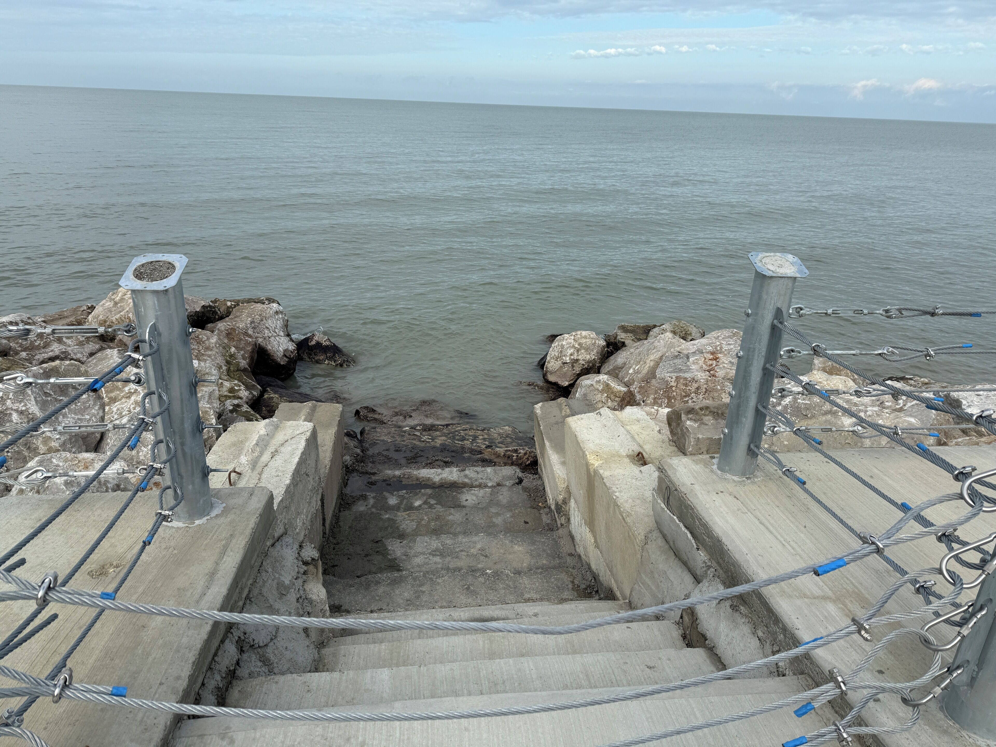 A concrete staircase leading to a rocky shoreline with a metal railing and a metal pole.