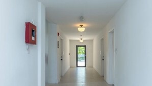 Un pasillo largo y estrecho con paredes blancas y suelo de tono claro. Hay puertas a ambos lados y un objeto rojo montado en la pared a la izquierda. El pasillo termina en una puerta.