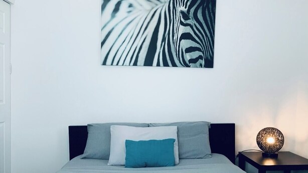 Una cama bien hecha con una manta gris y una toalla doblada, un cojín color aguamarina y una obra de arte con estampado de cebra en la pared.