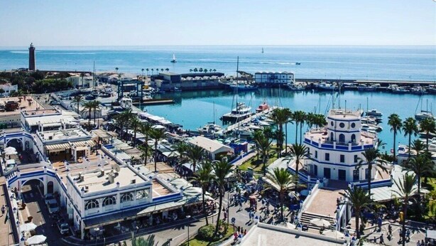Un port de plaisance situé sur la côte, avec des bateaux amarrés, un phare en arrière-plan et un mélange de bâtiments commerciaux et résidentiels.