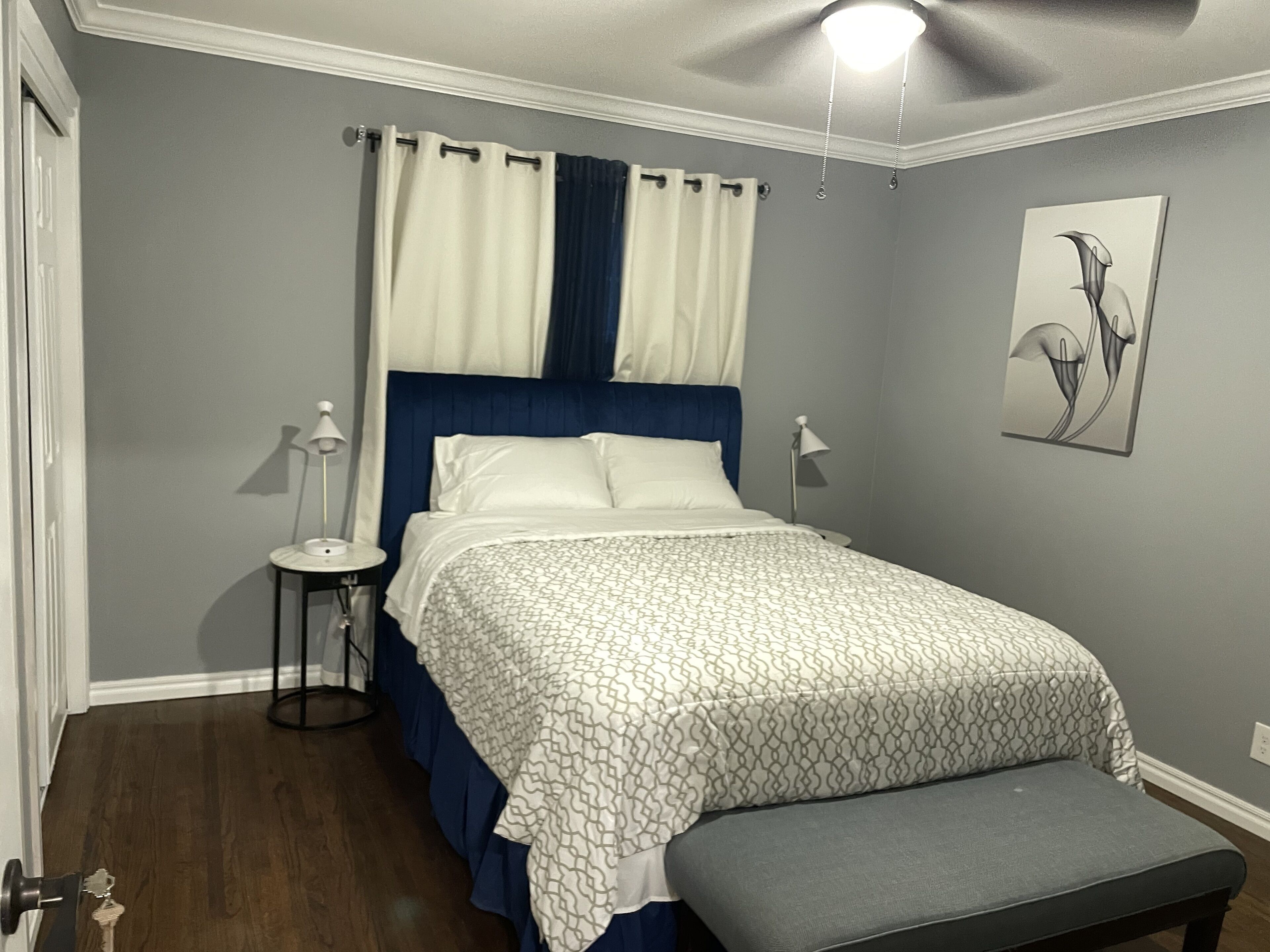 A bedroom with a bed, bedside lamps, a bench, a ceiling fan, and a wall-mounted artwork.