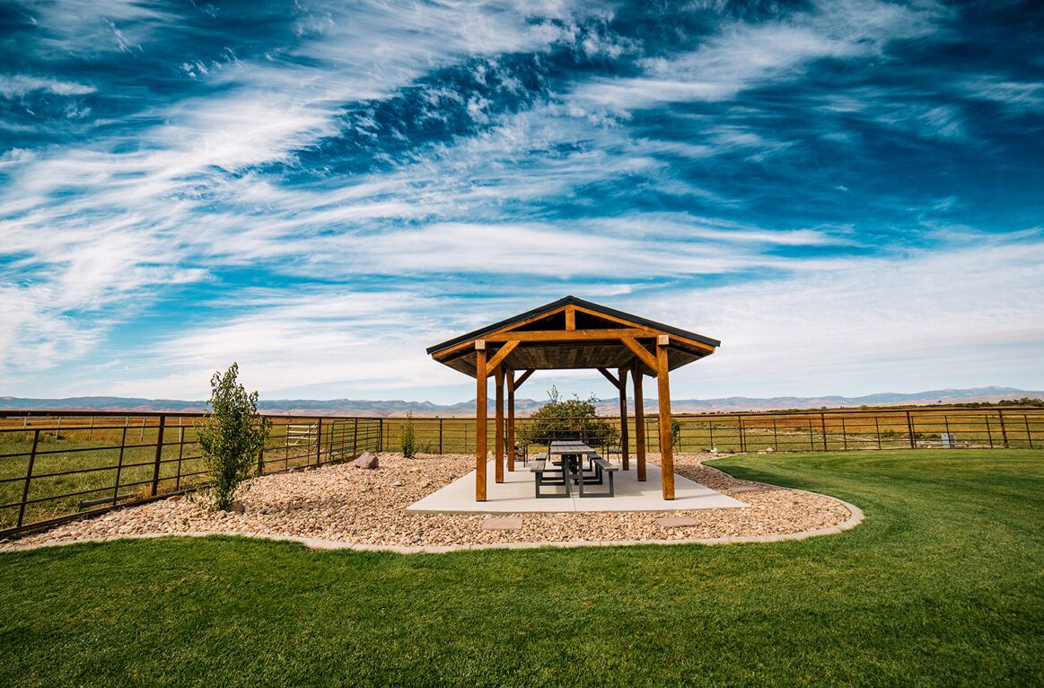 A wooden gazebo with picnic tables in a grassy field, surrounded by a fence and a clear sky.