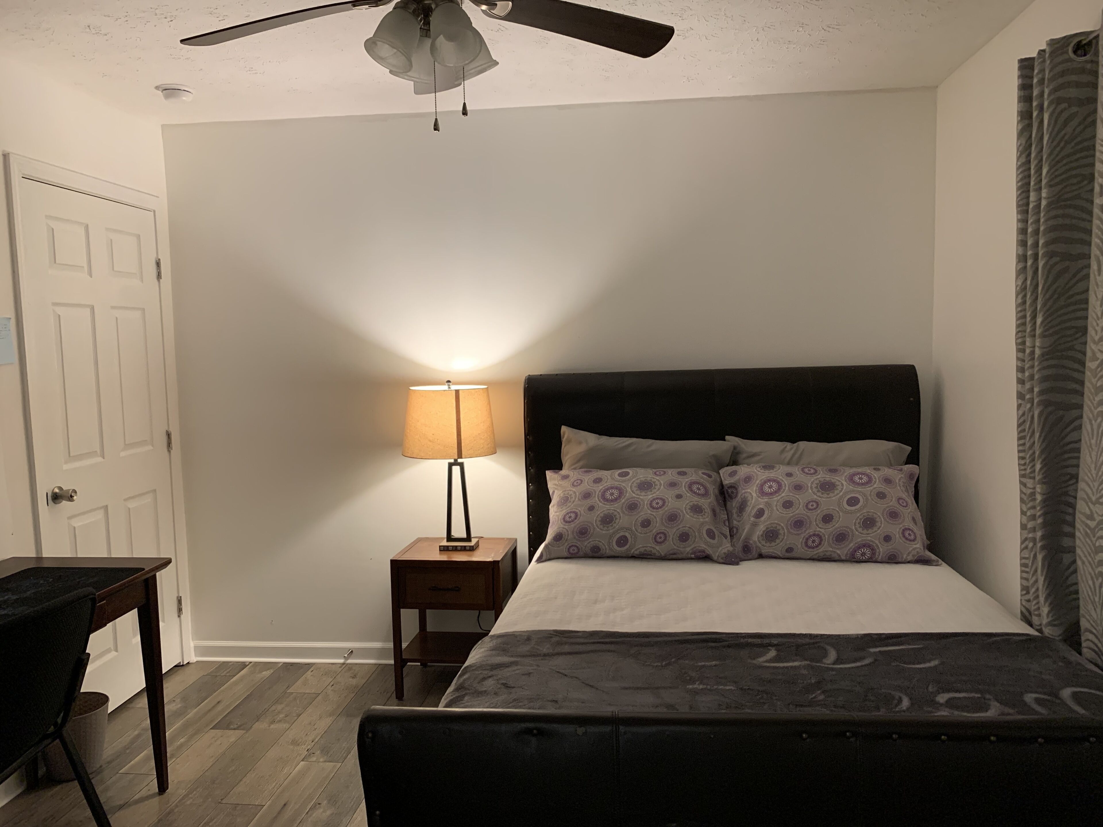 A bedroom with a bed, a nightstand, a lamp, a ceiling fan, and a door.
