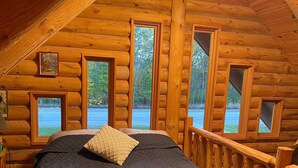 Un dormitorio tipo cabaña de troncos con una cama grande, varias ventanas y techo de madera.