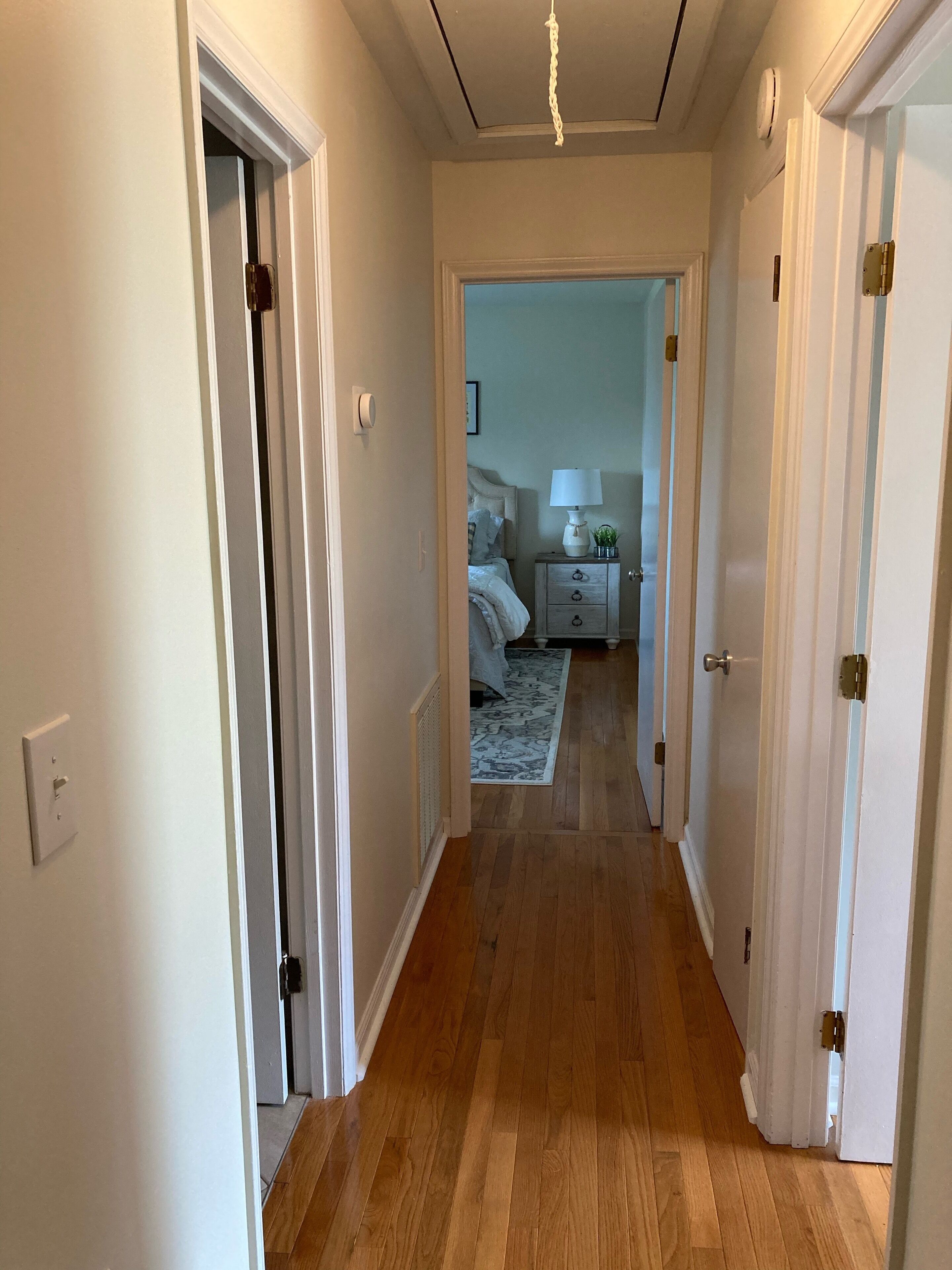 Un couloir au plancher en bois mène à une chambre à coucher qui contient un lit, une table de chevet, une lampe et une petite plante.