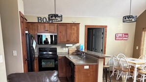 A modern kitchen with wooden cabinets, a black refrigerator, and a microwave. There is a dining area with a white table and chairs. A brown leather chair is in the foreground. - Waterpark passes included Bridges Bay Resort Arnold Park/Okoboji. (Arnolds Park)