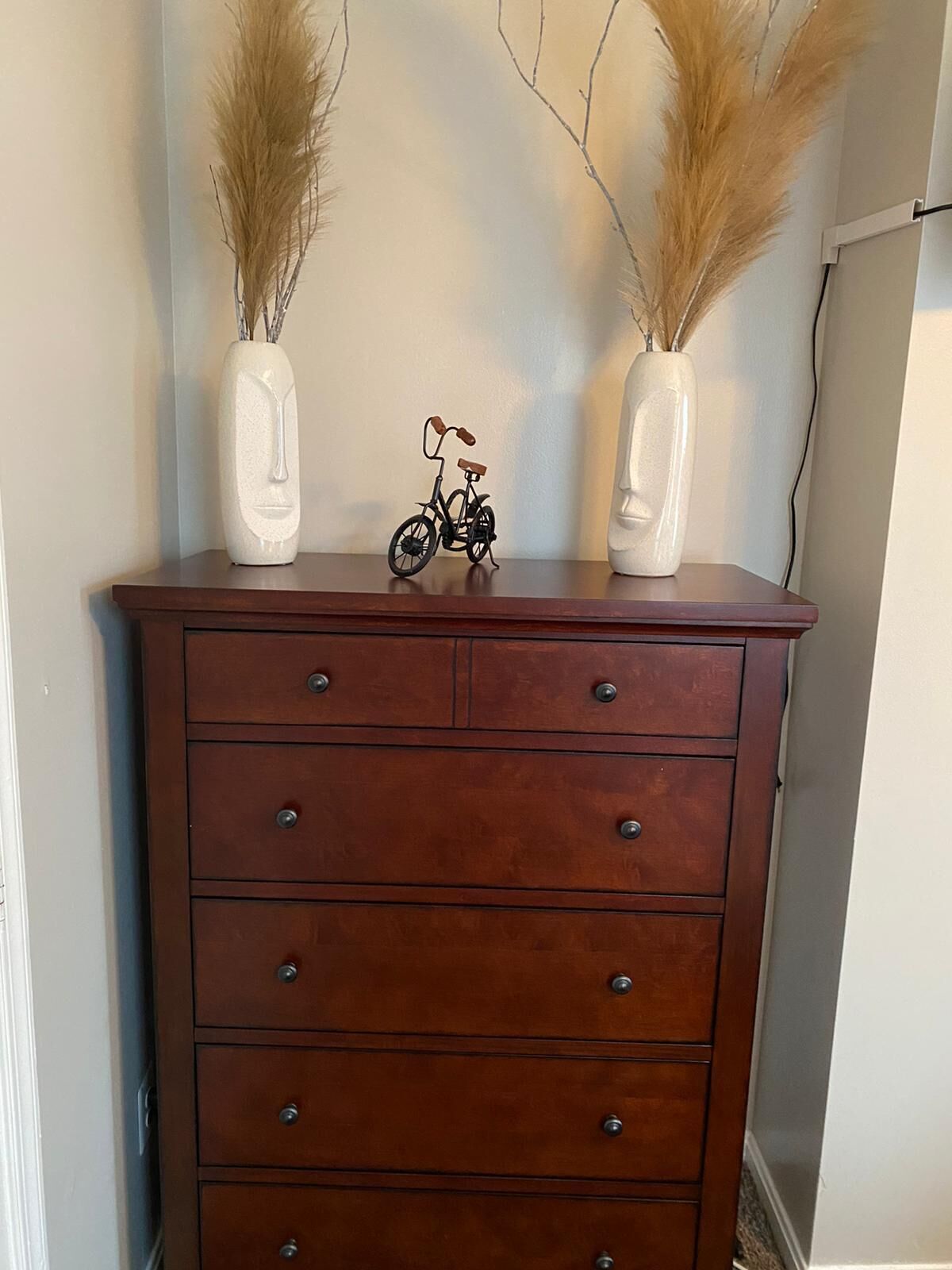 A wooden dresser with multiple drawers and two white vases with pampas grass on top.