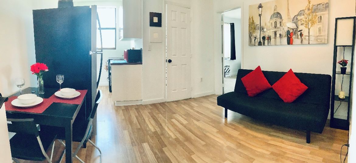A modern living room with a black sofa and red cushions, a dining area with a table set for two, and a hallway leading to a door.