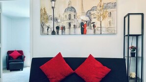 A living room with a black sofa, two red cushions, a painting of a street scene, and a black bookshelf with a plant. - Stylish 1 Brm Apartment in the Heart of Miami! #9 (Miami)