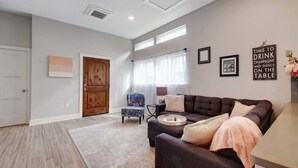 Un salon moderne avec un canapé marron foncé, un fauteuil à motifs et une petite table d’appoint. Il y a deux fenêtres avec des rideaux légers, une porte en bois et un mur orné de tableaux et d’une citation inspirante.