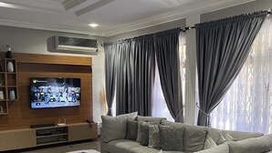 Uma sala de estar moderna com um sofá modular, uma TV de tela plana presa a uma parede de madeira e uma janela grande com cortinas cinzas.