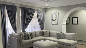 Uma sala de estar moderna com um sofá seccional, um banquinho combinando e uma grande janela com cortinas.
