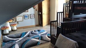 Een kamer met een houten vloer, een zwarte bank, een vloerkleed met een patroon, en een deur naar een andere ruimte.