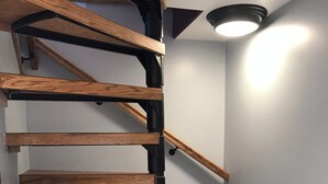 Un escalier en colimaçon avec des marches en bois et des rampes en métal noir, menant à un palier où se trouve un luminaire.
