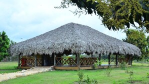 Una estructura con techo de paja, con una terraza de madera, rodeada de césped y árboles.