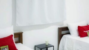 Habitación de hotel con dos camas, cada una con ropa de cama blanca y almohadas rojas. Hay una mesita negra con un objeto blanco encima y una cortina blanca al fondo.