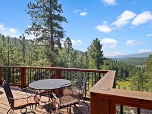 A wooden deck with a round table and chairs, overlooking a forested landscape with mountains in the distance. - Valley View Cabin, New Hot Tub, Close to Ski Lifts! (Angel Fire)