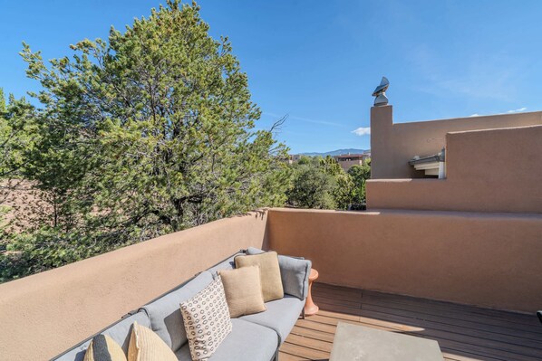 A rooftop terrace with a sofa and cushions, surrounded by trees and a clear sky.