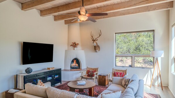 A cozy living room with a fireplace, a mounted deer head, a television, and a large window with a view of trees. - Spacious and Contemporary, Huge Primary Suite (Santa Fe)