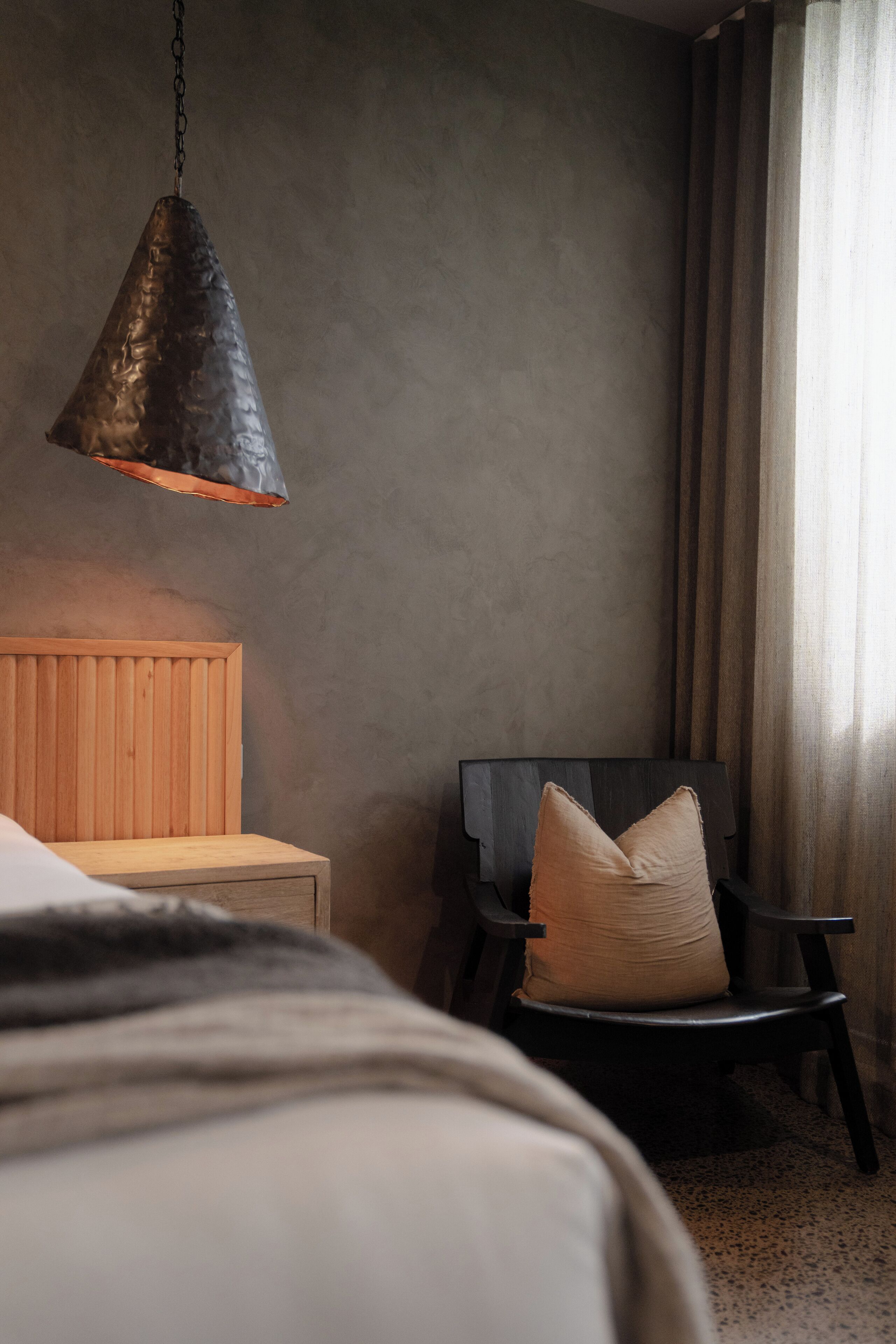 Ett sovrum med en sänggavel i trä, en svart stol med en kudde och en hängande lampa.
