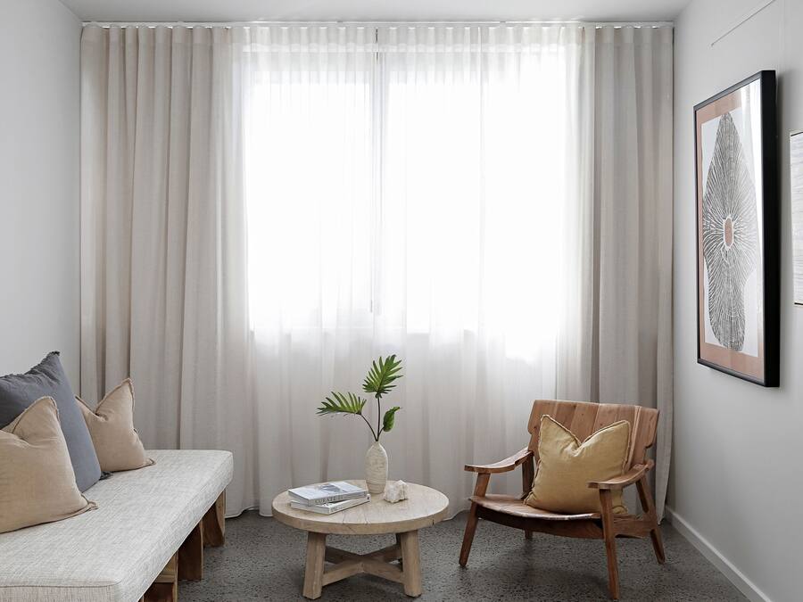 Habitación con sofá, una silla, una mesita con un florero y libros, y un ventanal con cortinas translúcidas.