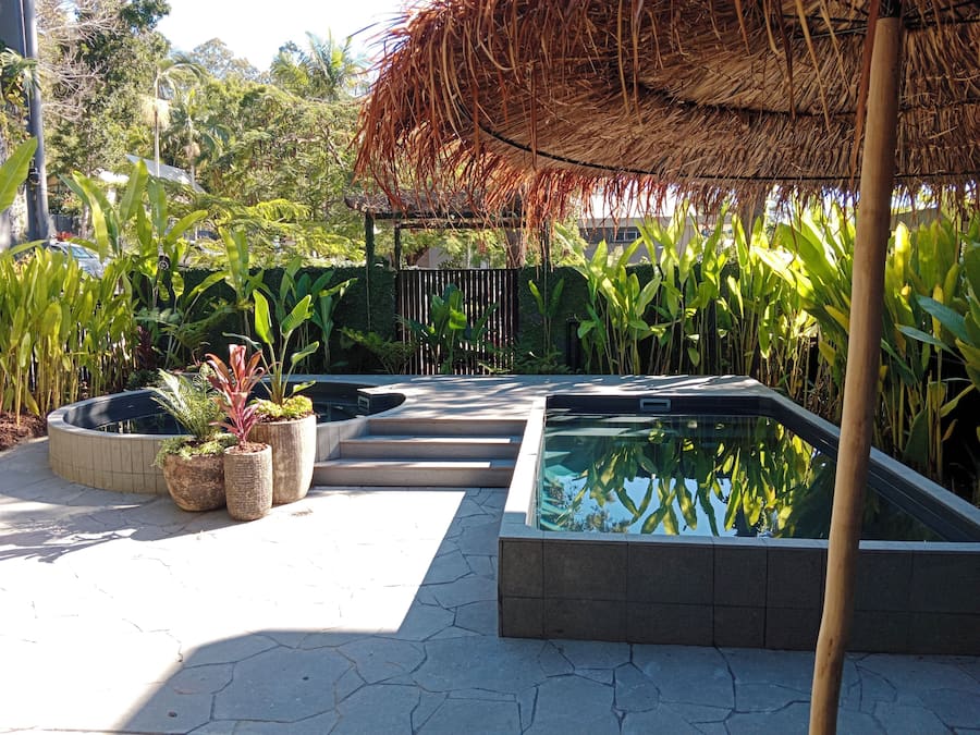 Ein Swimmingpool unter einem Reetdach, umgeben von üppigem Grün und einer gepflasterten Terrasse.