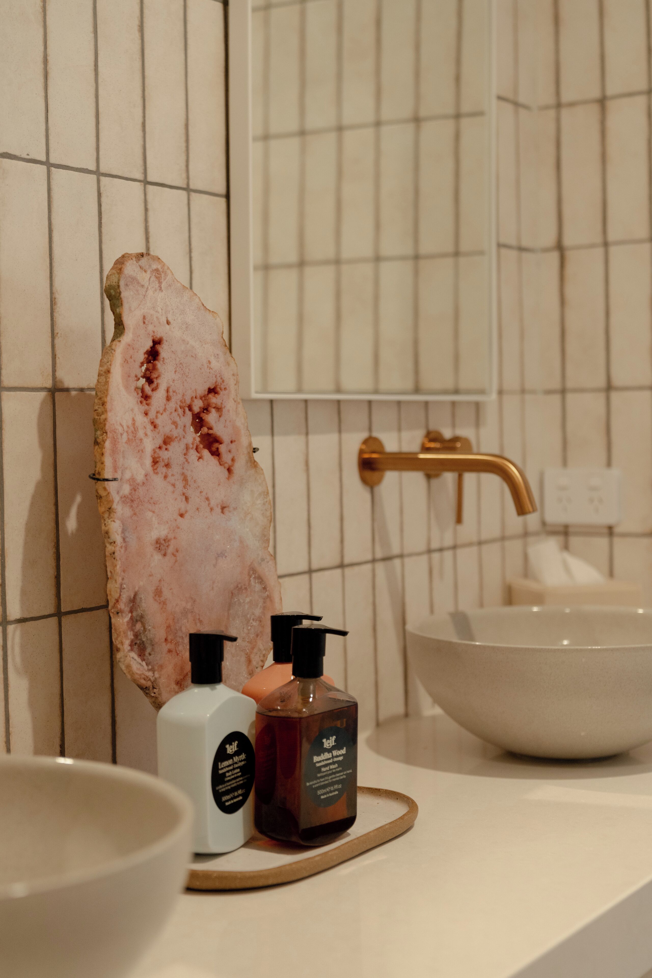 A bathroom with a large white basin, a gold faucet, and two bottles of soap on a wooden tray.
