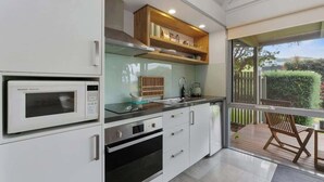 A modern kitchen with a built-in microwave, oven, and a wooden shelf above the sink. - Bay Beach Hut ~ Heated Floor ~ 50m Cafes (Apollo Bay)
