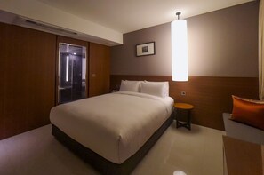 Una habitación de hotel con una cama grande, una mesita de noche, una silla, una lámpara en la pared y una puerta que da al baño.